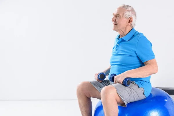 Uomo anziano con manubri seduto su palla fitness su sfondo grigio — Foto stock