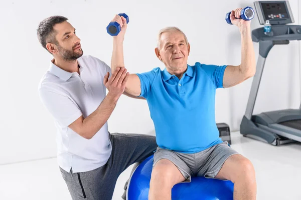 Terapeuta de reabilitação ajudando o homem idoso a se exercitar com halteres na bola de fitness em pano de fundo cinza — Fotografia de Stock