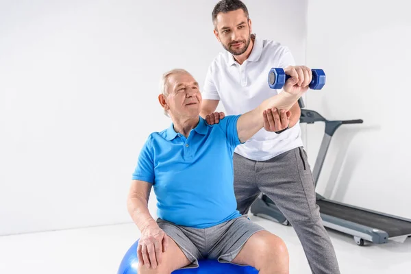 Portrait of rehabilitation therapist assisting senior man exercising with dumbbell on grey backdrop — Stock Photo