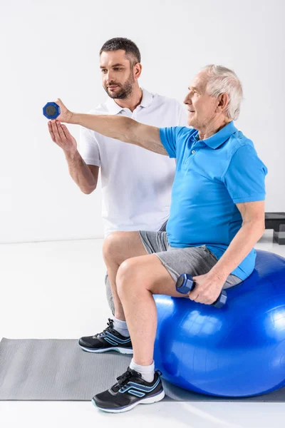 Реабілітаційний терапевт, який допомагає старшому чоловікові тренуватися з гантелями на фітнес-кулі — стокове фото