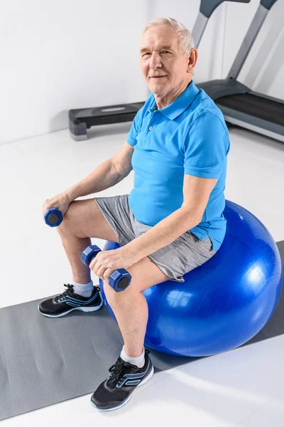 High angle view of smiling senior man with dumbbells sitting on fitness ball on grey backdrop — Stock Photo
