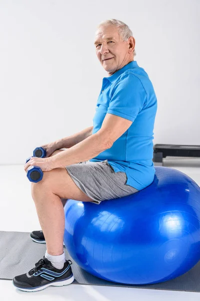 Vista lateral do homem sênior com halteres sentados na bola de fitness em fundo cinza — Fotografia de Stock