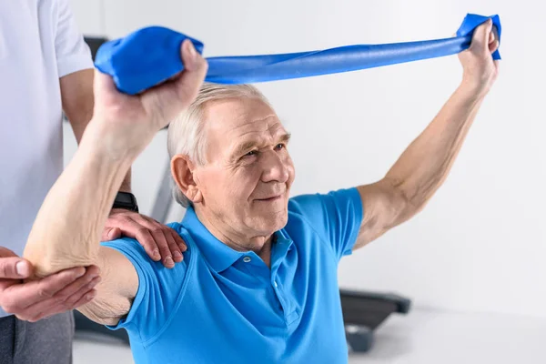 Cropped shot of rehabilitation therapist assisting senior man exercising with rubber tape — Stock Photo