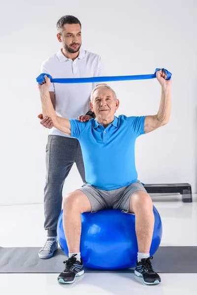 Terapeuta de reabilitação ajudando o homem idoso a se exercitar com fita de borracha na bola de fitness — Fotografia de Stock