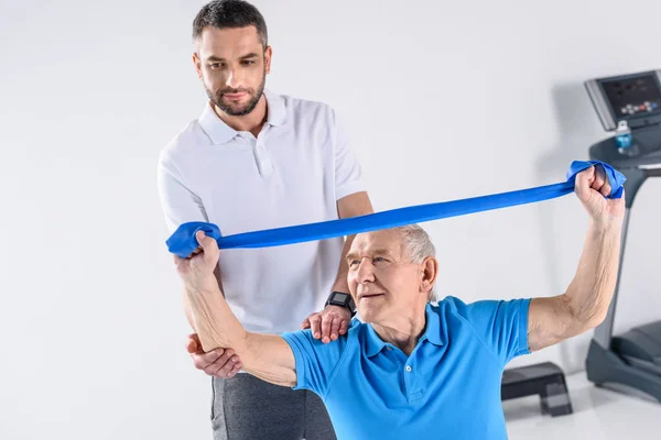 Rehabilitation therapist assisting senior man exercising with rubber tape — Stock Photo