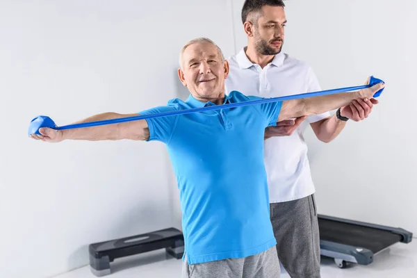 Terapeuta de rehabilitación que ayuda a hombre mayor sonriente a hacer ejercicio con cinta de goma - foto de stock
