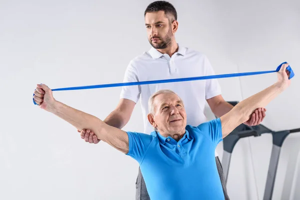 Retrato del terapeuta de rehabilitación que ayuda al hombre mayor a hacer ejercicio con cinta de goma - foto de stock