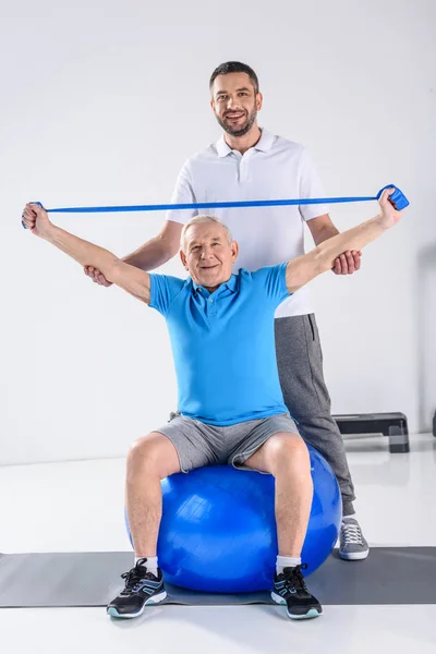 Terapeuta de reabilitação assistindo sorridente homem sênior exercitando com fita de borracha na bola de fitness — Fotografia de Stock