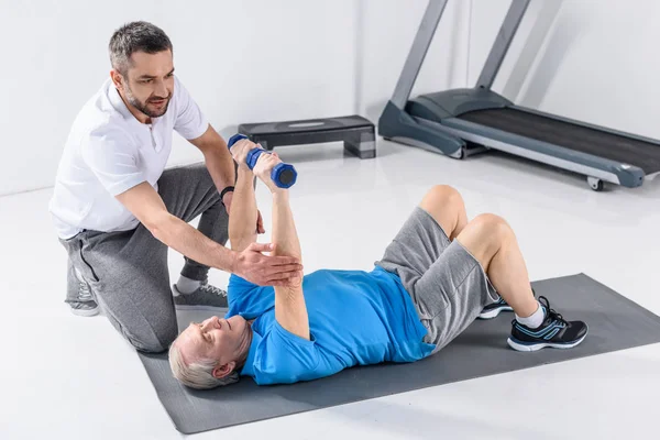 Rehabilitation therapist assisting senior man exercising with dumbbells on mat — Stock Photo