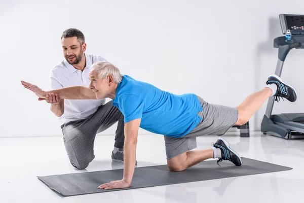 Rehabilitation therapist helping smiling senior man exercising on mat — Stock Photo