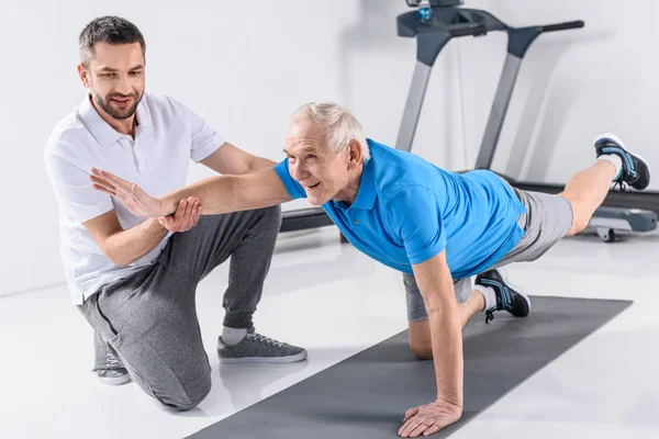 Rehabilitation therapist helping smiling senior man exercising on mat — Stock Photo