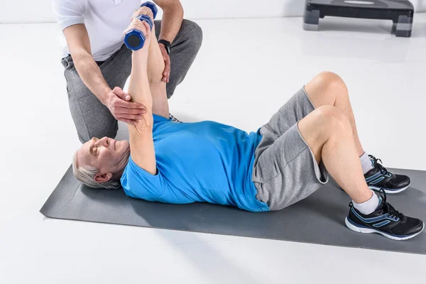 Vista parcial del terapeuta de rehabilitación que ayuda al hombre mayor a hacer ejercicio con pesas en la esterilla — Stock Photo