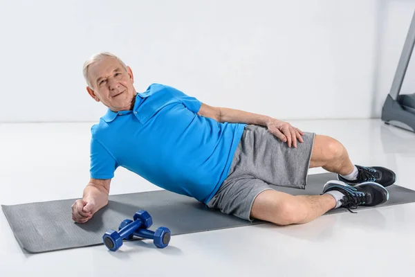 Smiling senior man resting on mat with dumbbells near by on grey backdrop — Stock Photo