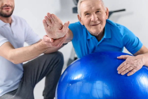 Vista parcial del terapeuta de rehabilitación que ayuda al hombre mayor sonriente que hace ejercicio en la pelota de fitness - foto de stock