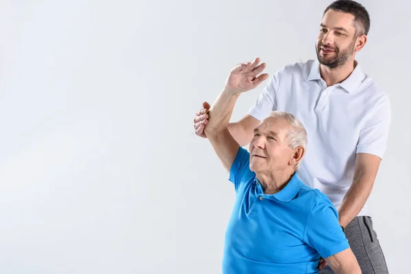 Retrato del terapeuta de rehabilitación ayudando al hombre mayor a estirarse aislado en gris - foto de stock