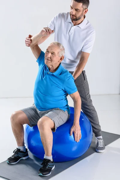 Reha-Therapeutin hilft Senioren beim Training auf Fitnessball vor grauem Hintergrund — Stockfoto