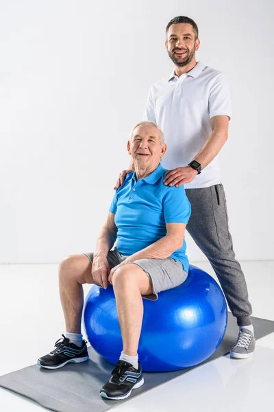 Terapeuta de rehabilitación sonriente y hombre mayor en la pelota de fitness mirando a la cámara en el fondo gris - foto de stock