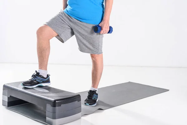 Partial view of senior man with dumbbells exercising on stepper isolated on grey — Stock Photo