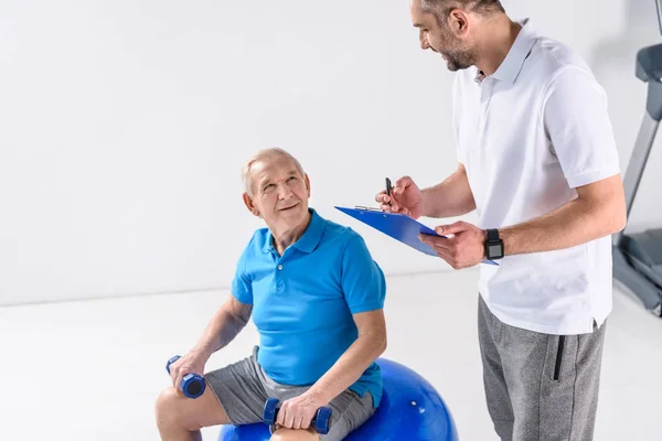 Reha-Therapeut mit Notizblock hilft Senioren beim Training mit Hanteln vor grauem Hintergrund — Stockfoto