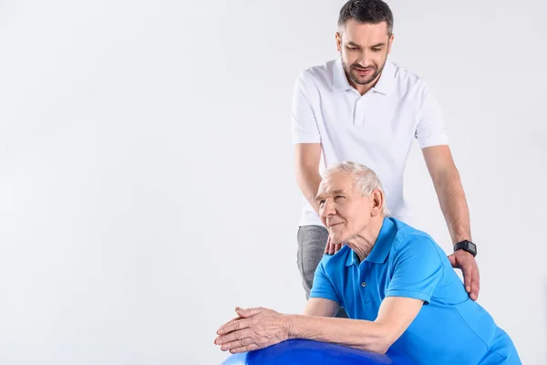 Portrait of rehabilitation therapist making massage to senior man on fitness ball isolated on grey — Stock Photo