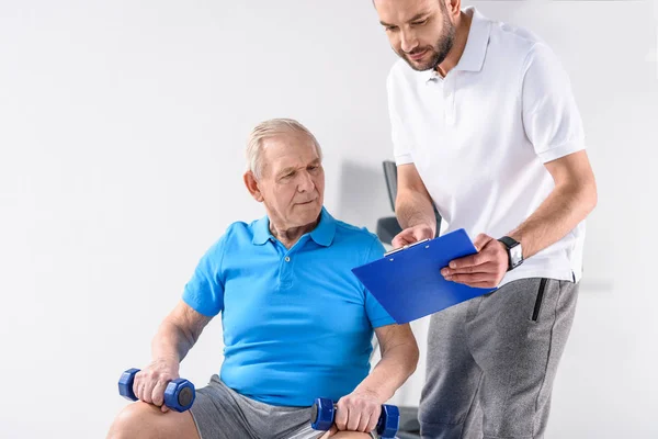 Terapeuta di riabilitazione con blocco note che assiste l'uomo anziano nell'esercizio con manubri su sfondo grigio — Foto stock