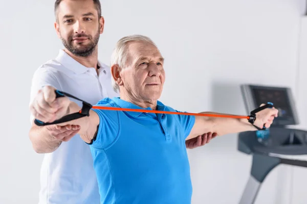 Thérapeute de réadaptation souriant aider l'homme âgé exercice avec du ruban de caoutchouc — Photo de stock