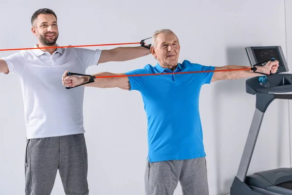 Smiling rehabilitation therapist and senior man exercising with rubber tapes — Stock Photo