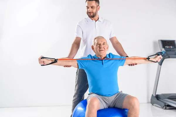 Terapeuta de rehabilitación que ayuda a un hombre mayor a hacer ejercicio con cinta de goma en la pelota de fitness con fondo gris - foto de stock