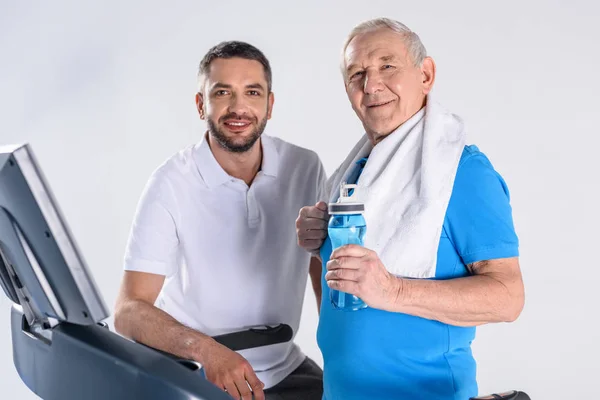 Портрет улыбающегося реабилитационного терапевта и пожилого человека со спортивной бутылкой воды на беговой дорожке, изолированной на сером — стоковое фото