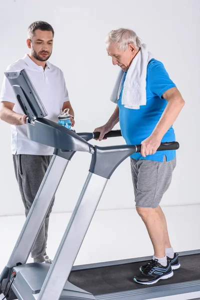 Terapeuta de reabilitação auxiliando o homem idoso com toalha de exercício em esteira isolada em cinza — Fotografia de Stock
