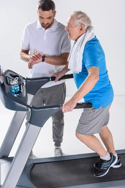 Rehabilitation therapist checking time while assisting senior man exercising on treadmill isolated on grey — Stock Photo
