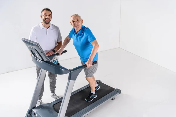 Vista de ángulo alto del terapeuta de rehabilitación que ayuda al hombre mayor a hacer ejercicio en la cinta de correr - foto de stock