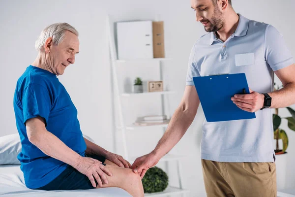 Vista lateral del terapeuta de rehabilitación con bloc de notas comprobando la rodilla del hombre mayor en la mesa de masaje - foto de stock