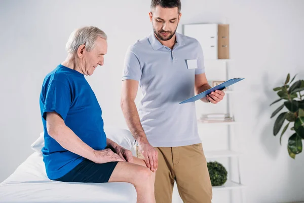 Terapeuta de rehabilitación con bloc de notas comprobando la rodilla del hombre mayor en la mesa de masaje - foto de stock