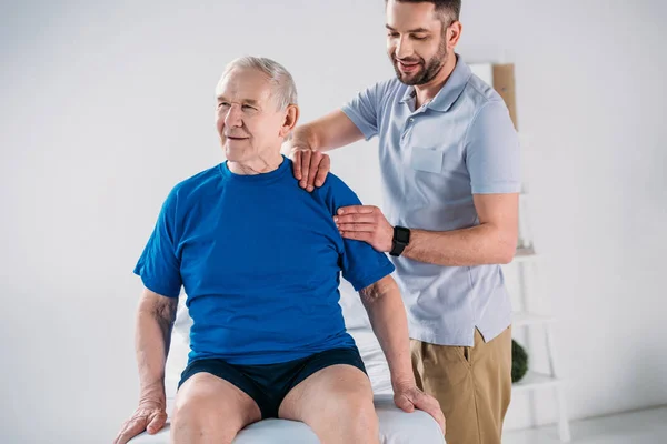 Retrato del fisioterapeuta haciendo masaje al hombre mayor en la mesa de masaje - foto de stock