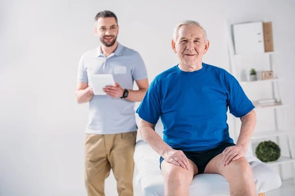 Вибірковий фокус реабілітаційного терапевта з планшетом і усміхненим старшим чоловіком на масажному столі — стокове фото