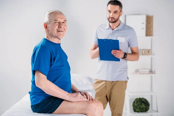 Focus selettivo del terapeuta riabilitativo con blocco note e uomo anziano sul tavolo da massaggio — Foto stock