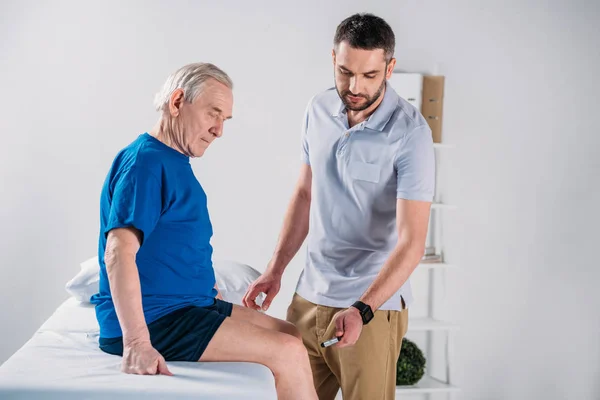 Terapeuta de rehabilitación con martillo reflexivo revisando la rodilla del hombre mayor en la mesa de masaje - foto de stock