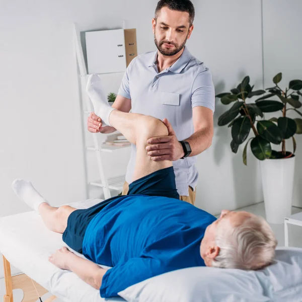 Terapeuta riabilitativo concentrato massaggiare senior mans gamba sul tavolo da massaggio — Foto stock