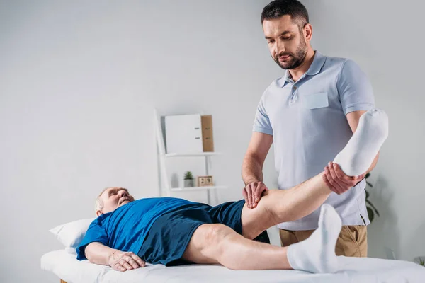 Terapeuta de reabilitação focada massageando perna de homem sênior na mesa de massagem — Fotografia de Stock