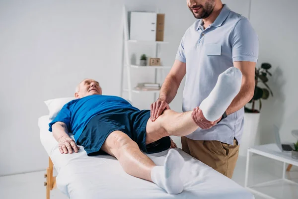 Visão parcial do fisioterapeuta massageando a perna do homem sênior na mesa de massagem — Fotografia de Stock