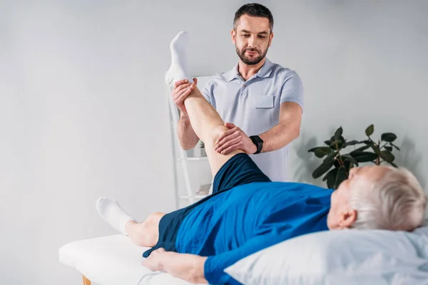 Portrait of rehabilitation therapist massaging senior mans leg on massage table — Stock Photo