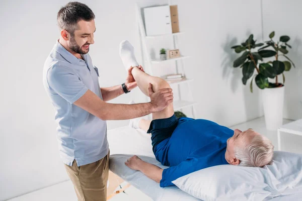 Fisioterapeuta sonriente masajeando la pierna del hombre mayor en la mesa de masaje - foto de stock