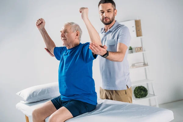 Retrato del fisioterapeuta enfocado haciendo masaje al hombre mayor en la mesa de masaje - foto de stock