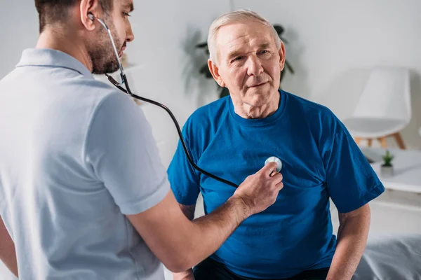 Реабілітаційний терапевт зі стетоскопом, який перевіряє серцебиття старшого чоловіка — стокове фото
