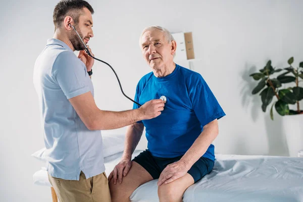 Terapeuta de rehabilitación con estetoscopio revisando los latidos del corazón de los hombres mayores - foto de stock