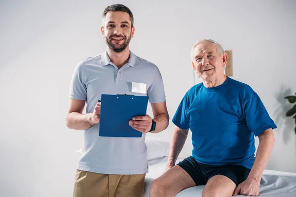 Retrato del terapeuta de rehabilitación con bloc de notas y hombre mayor sonriente en la mesa de masaje - foto de stock