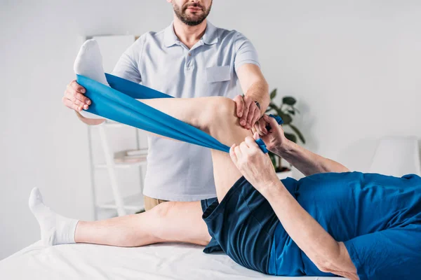 Cropped shot of rehabilitation therapist assisting senior man exercising with rubber tape on massage table — Stock Photo