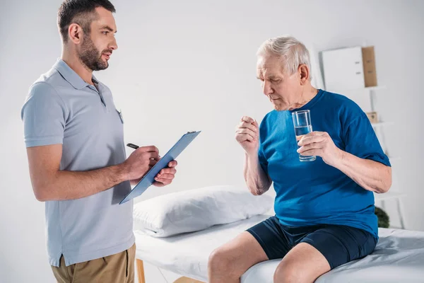 Vue latérale du thérapeute de réadaptation avec bloc-notes et homme âgé avec verre d'eau et pilule sur table de massage — Photo de stock