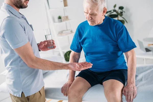 Tiro recortado de terapeuta de rehabilitación dando píldora al hombre mayor en la mesa de masaje - foto de stock